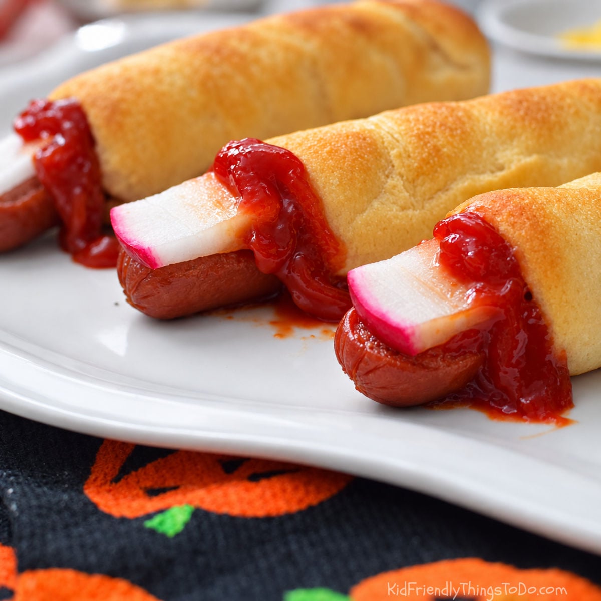 alt text Halloween hot dog fingers wrapped in golden dough with radish fingernails and ketchup, served on a white platter.