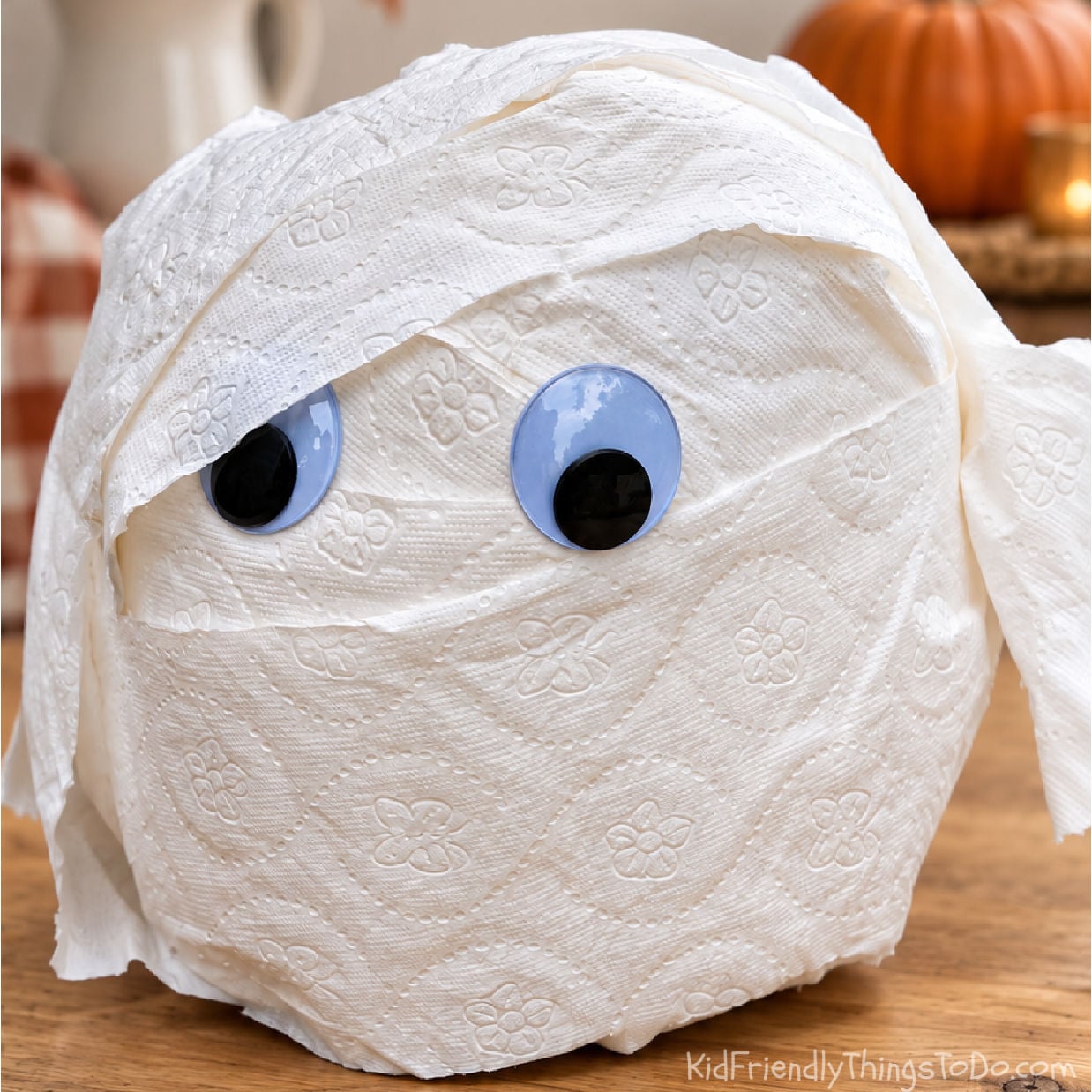 Toilet paper mummy prize ball with googly eyes on a wooden table, with softly blurred fall decorations behind it.
