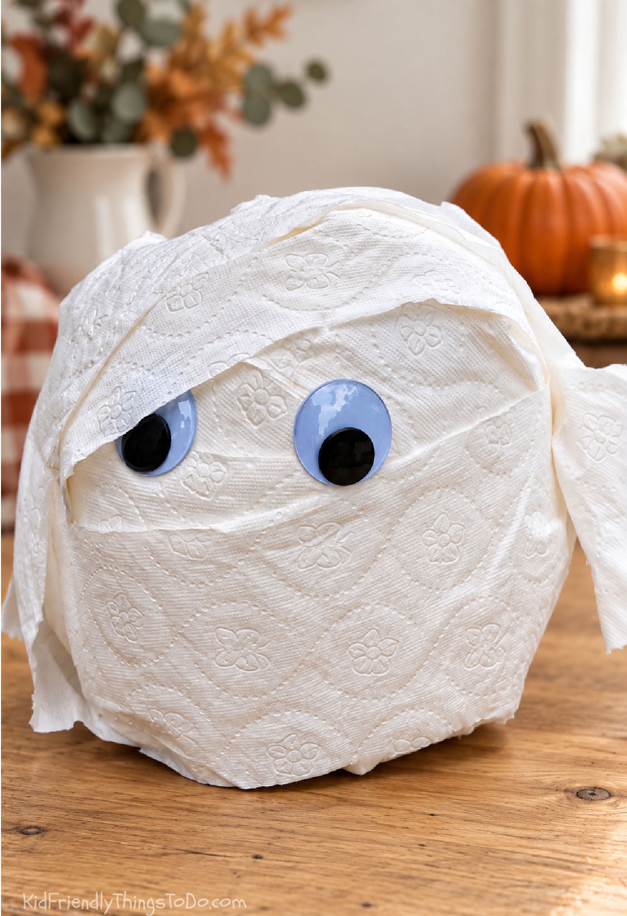 Toilet paper mummy prize ball with googly eyes on a wooden table, with softly blurred fall decorations behind it.