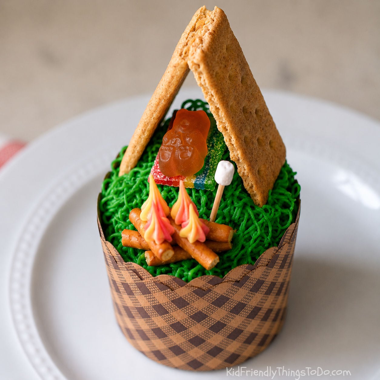 Campfire cupcakes decorated with green grass frosting, a graham cracker tent, candy campfire, marshmallow, and sleeping bag for a fun camping dessert idea for kids