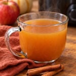 Warm mulled apple cider in a clear glass mug with cinnamon sticks on a wooden table and a slow cooker in the background.