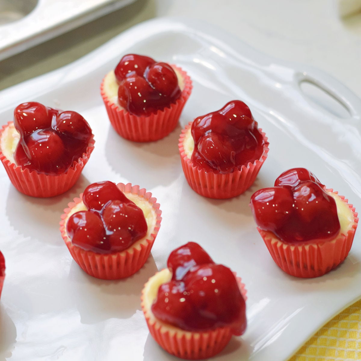 Mini cheesecake bites topped with cherry pie filling in red cupcake liners on a white serving platter.