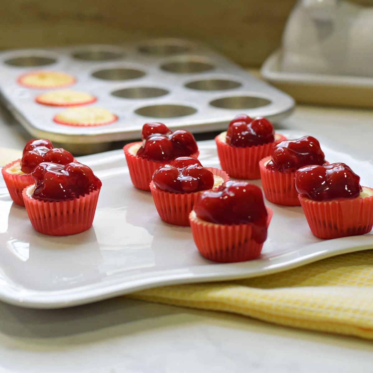 Mini cheesecake bites topped with cherry pie filling in red liners on a white serving platter.
