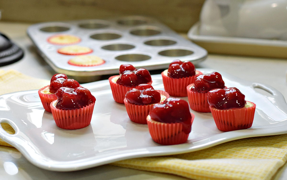 Mini cheesecake bites topped with cherry pie filling in red liners on a white serving platter.