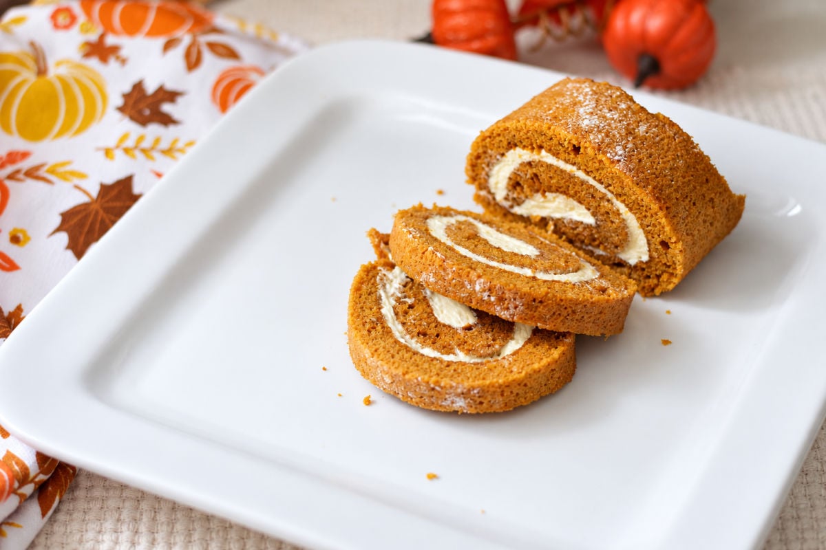 Two overlapping slices of pumpkin roll with cream cheese filling beside the remaining roll on a white platter.