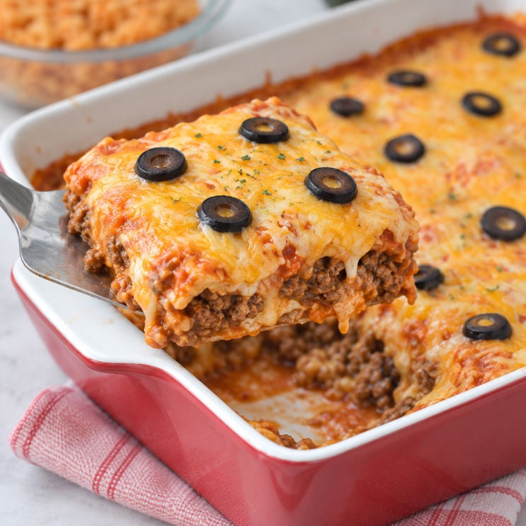 Beef enchilada casserole serving lifted from a red baking dish, showing ground beef and tortilla layers topped with melted cheese, red sauce, and sliced black olives.