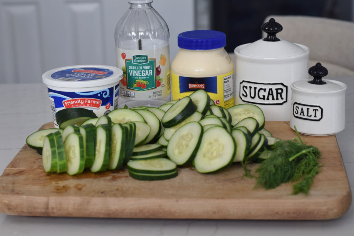 Creamy cucumber salad ingredients: sliced cucumbers, fresh dill, sour cream, mayonnaise, white vinegar, sugar, and salt arranged on a wooden cutting board.