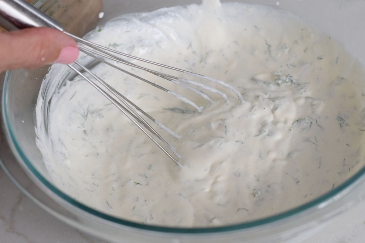 Whisking sour cream, mayonnaise, and dill dressing for creamy cucumber salad in a glass mixing bowl.