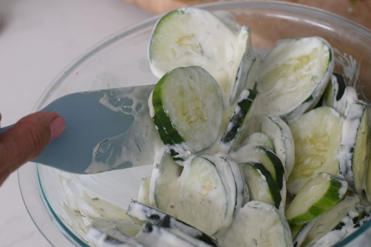 Folding sliced cucumbers into creamy dill dressing with a blue spatula in a glass mixing bowl.