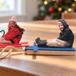 Two handmade sled ornaments with cutout photos of children sitting on red and blue sleds, displayed on a wooden table.