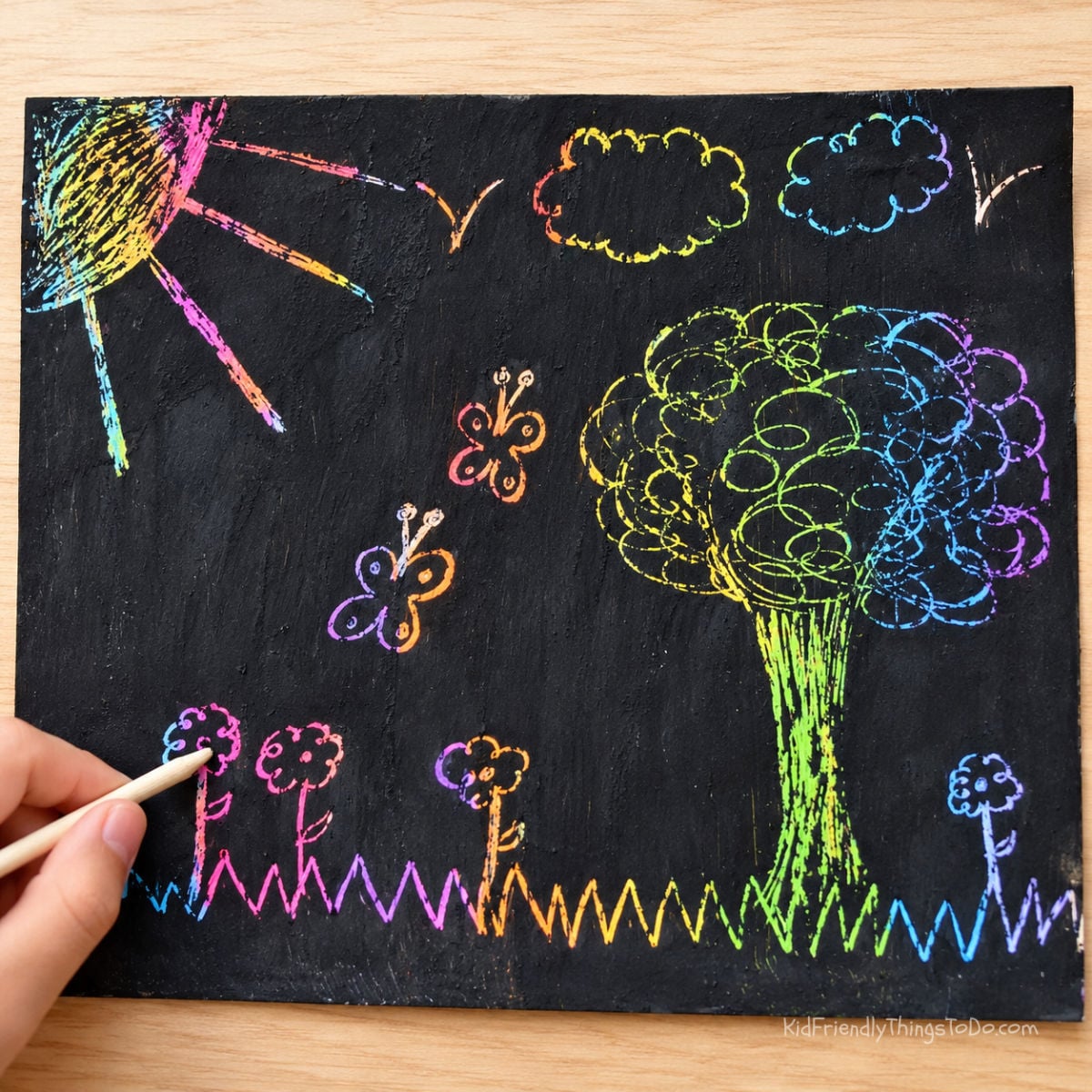 Rainbow scratch art for kids made with colorful crayons and black paint, featuring a hand scratching a design to reveal bright colors underneath.