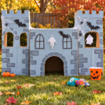 Cardboard haunted castle bean bag toss with gray painted walls, cutout targets, paper bats, cobwebs, and hanging ghosts, set on grass with colorful beanbags and a pumpkin bucket.