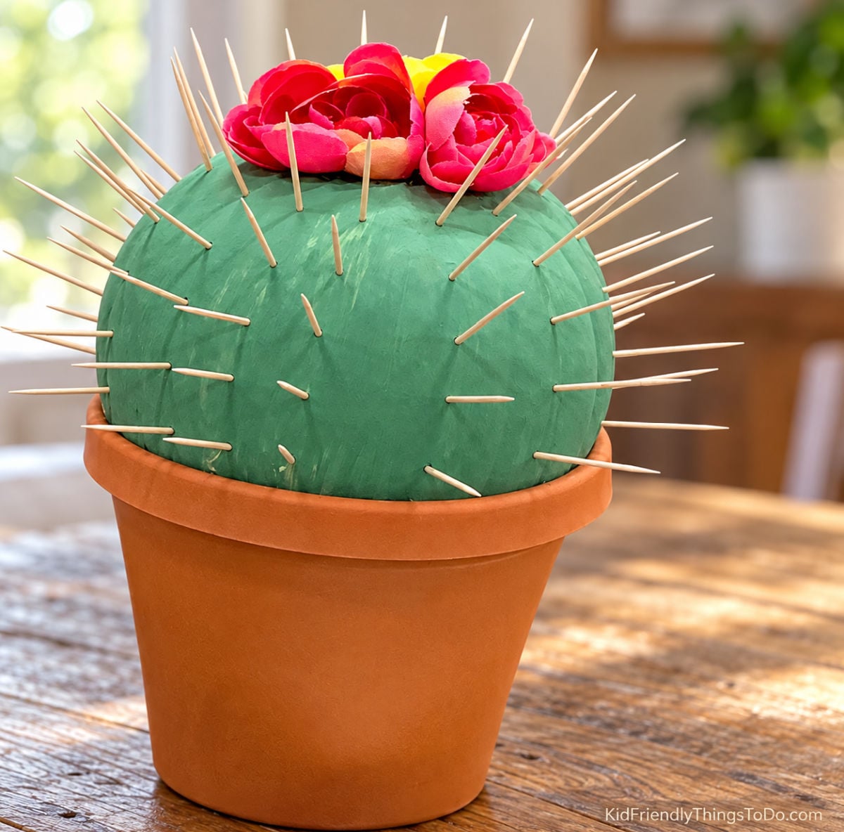 Green cactus pumpkin with wooden toothpick spines and bright pink flowers, displayed in a terracotta-colored flowerpot.