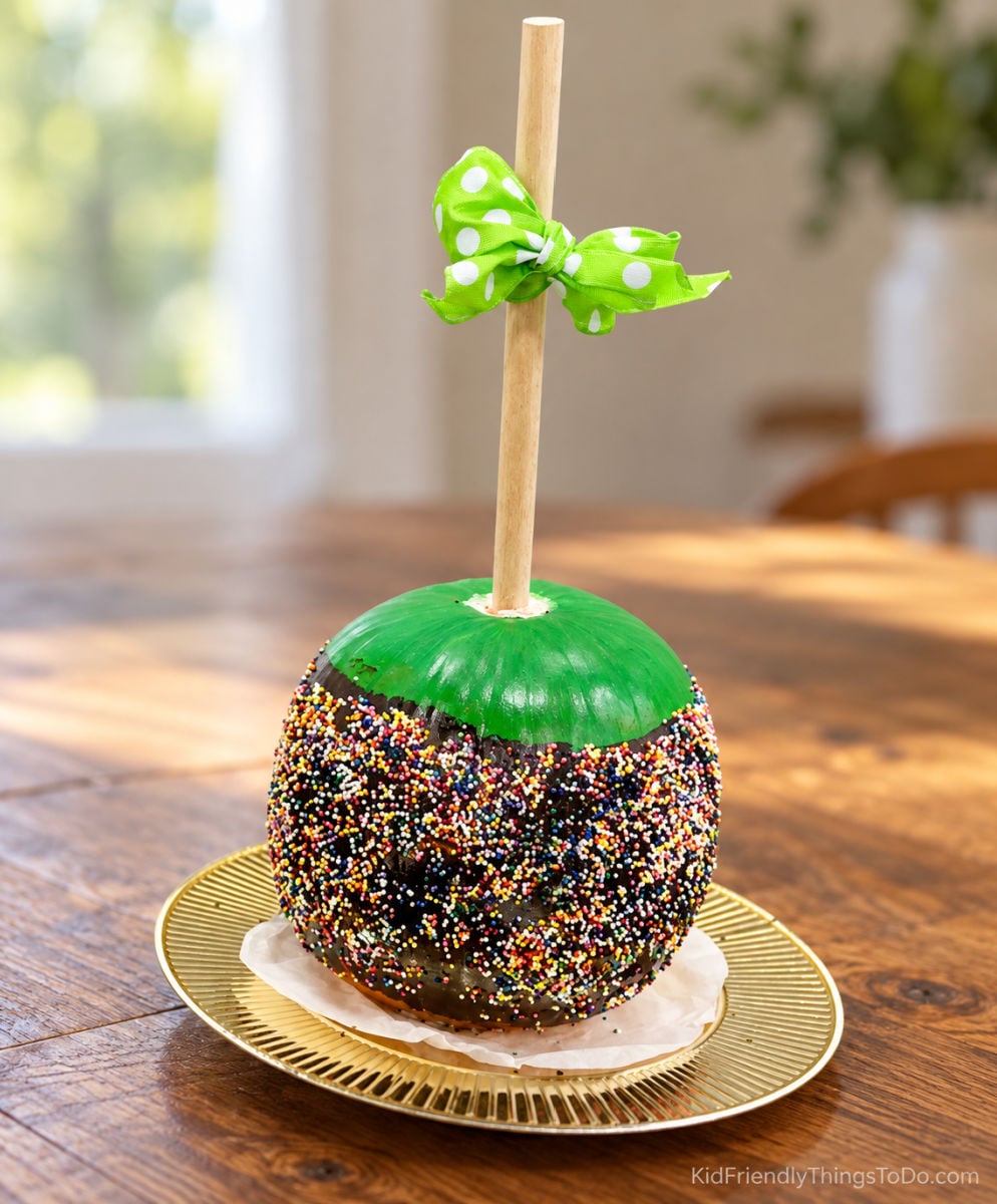 Candy apple pumpkin with a green top, sprinkle-covered brown sides, and a wooden stick tied with a green polka-dot bow.