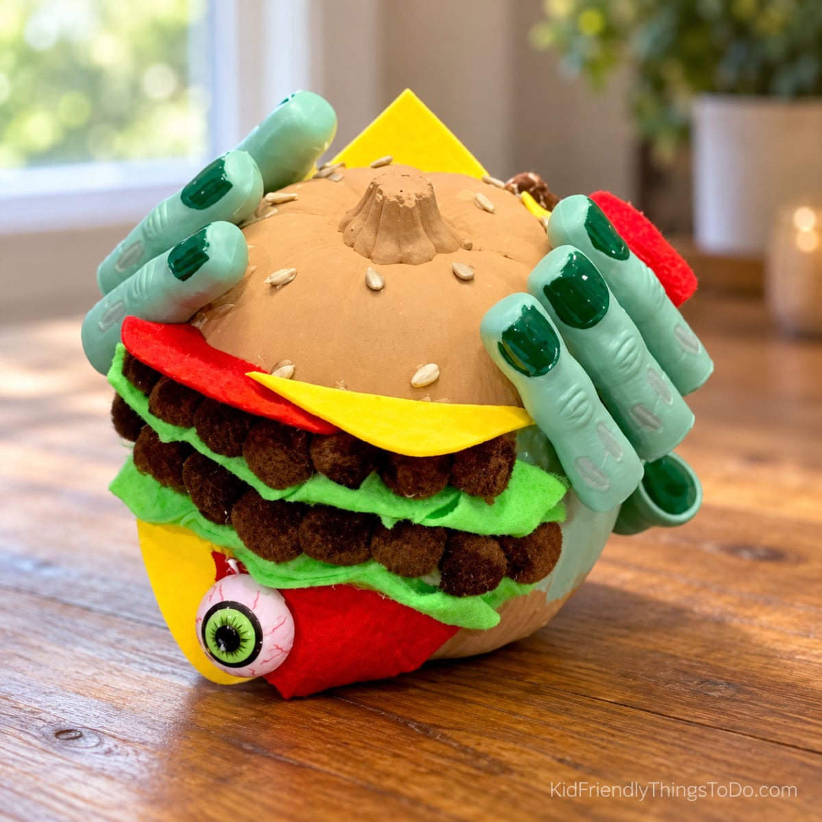 Monster cheeseburger pumpkin with brown pom-pom patties, green lettuce, yellow cheese, toy monster fingers, and a dangling eyeball.