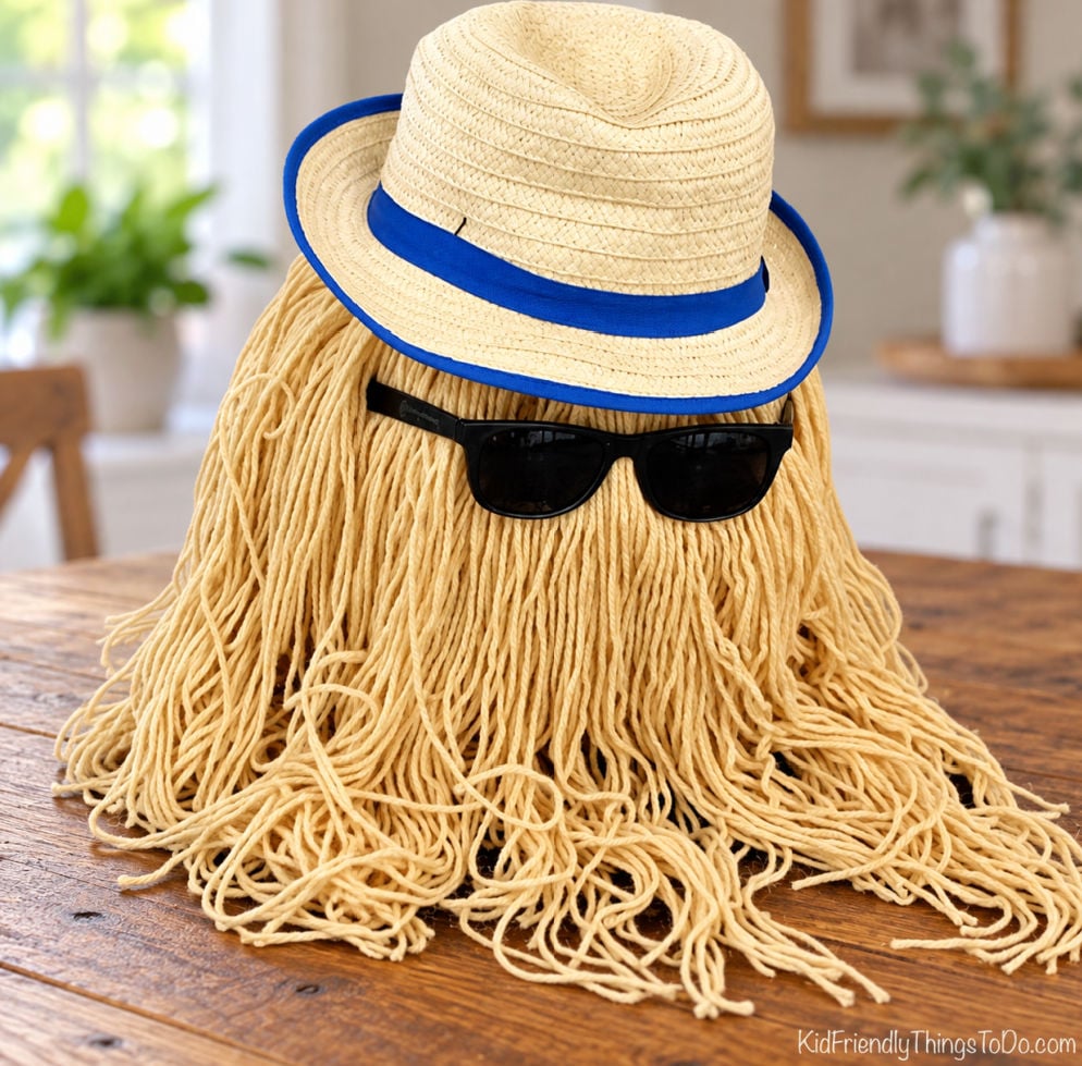 No-carve Cousin Itt pumpkin covered in long tan yarn, wearing black sunglasses and a cream hat with blue trim.