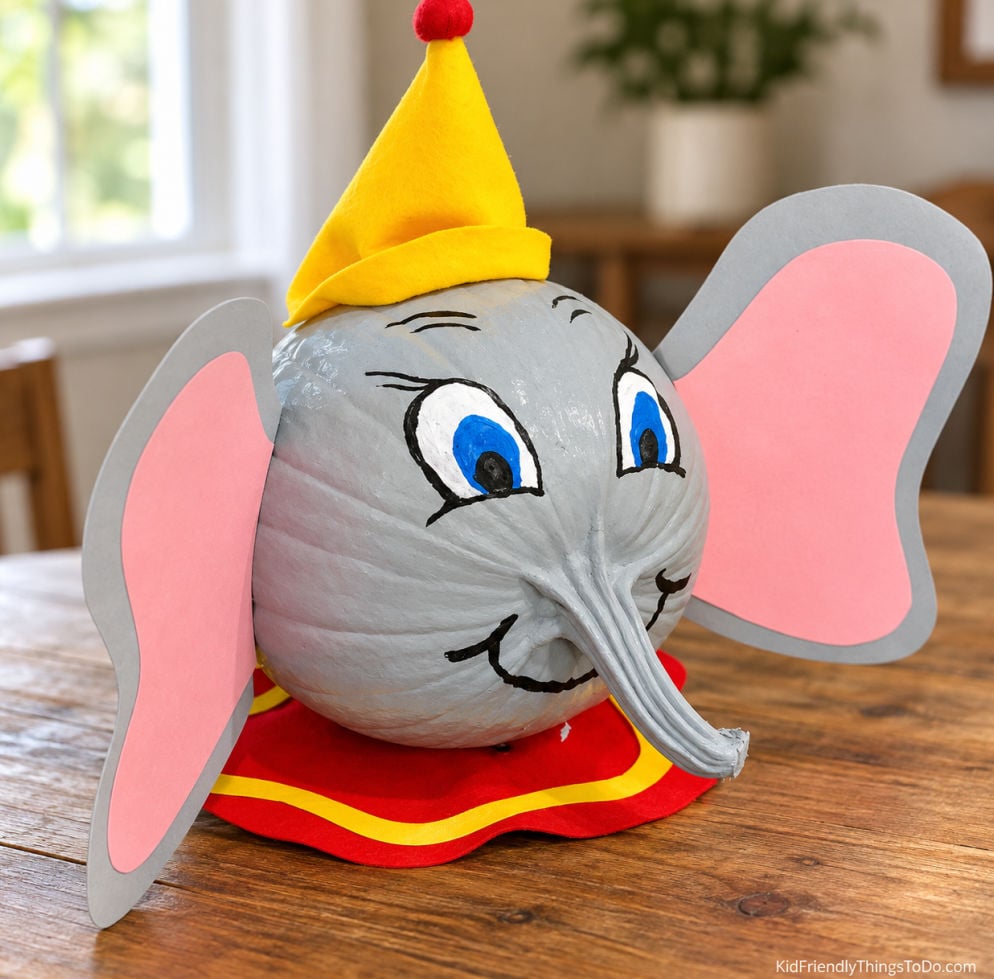 No-carve Dumbo pumpkin painted gray with large pink-lined ears, blue eyes, a curved trunk, and a yellow hat with a red pompom.
