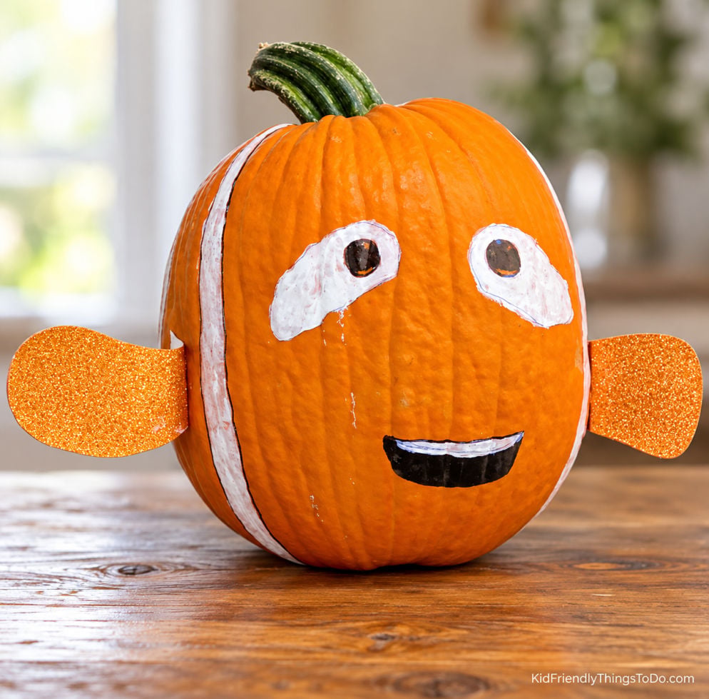 No-carve Nemo pumpkin with painted white stripes, a smiling face, and orange glitter fins, displayed on a wooden table.