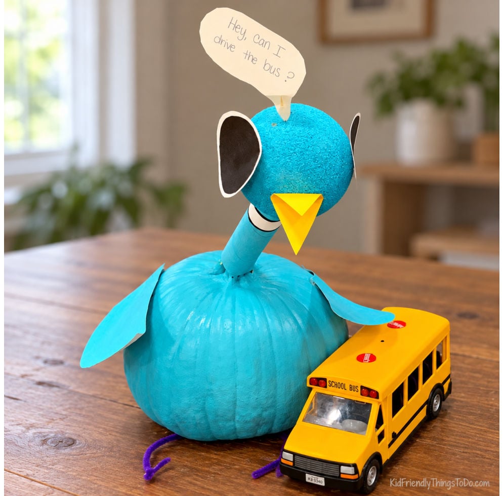 Turquoise no-carve pigeon pumpkin with a yellow beak beside a toy school bus and a speech bubble reading “Hey, can I drive the bus?