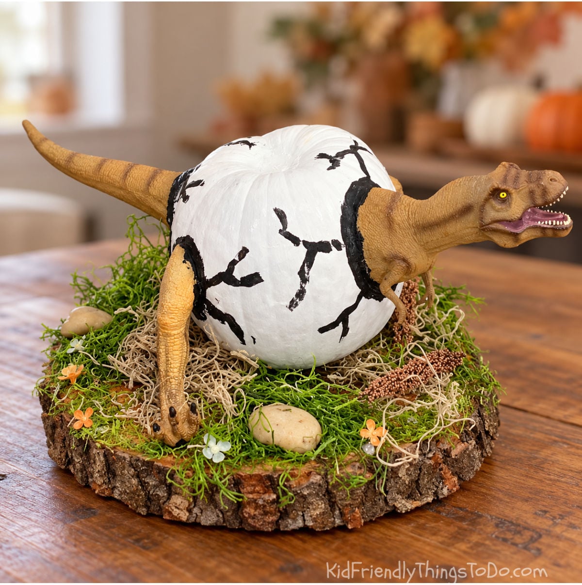 T. rex emerging from a white pumpkin painted like a cracked dinosaur egg, displayed on a moss-covered wood slice.