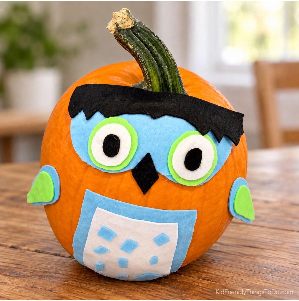 No-carve owl pumpkin decorated with blue, green, white, and black felt, large round eyes, and a patterned belly.