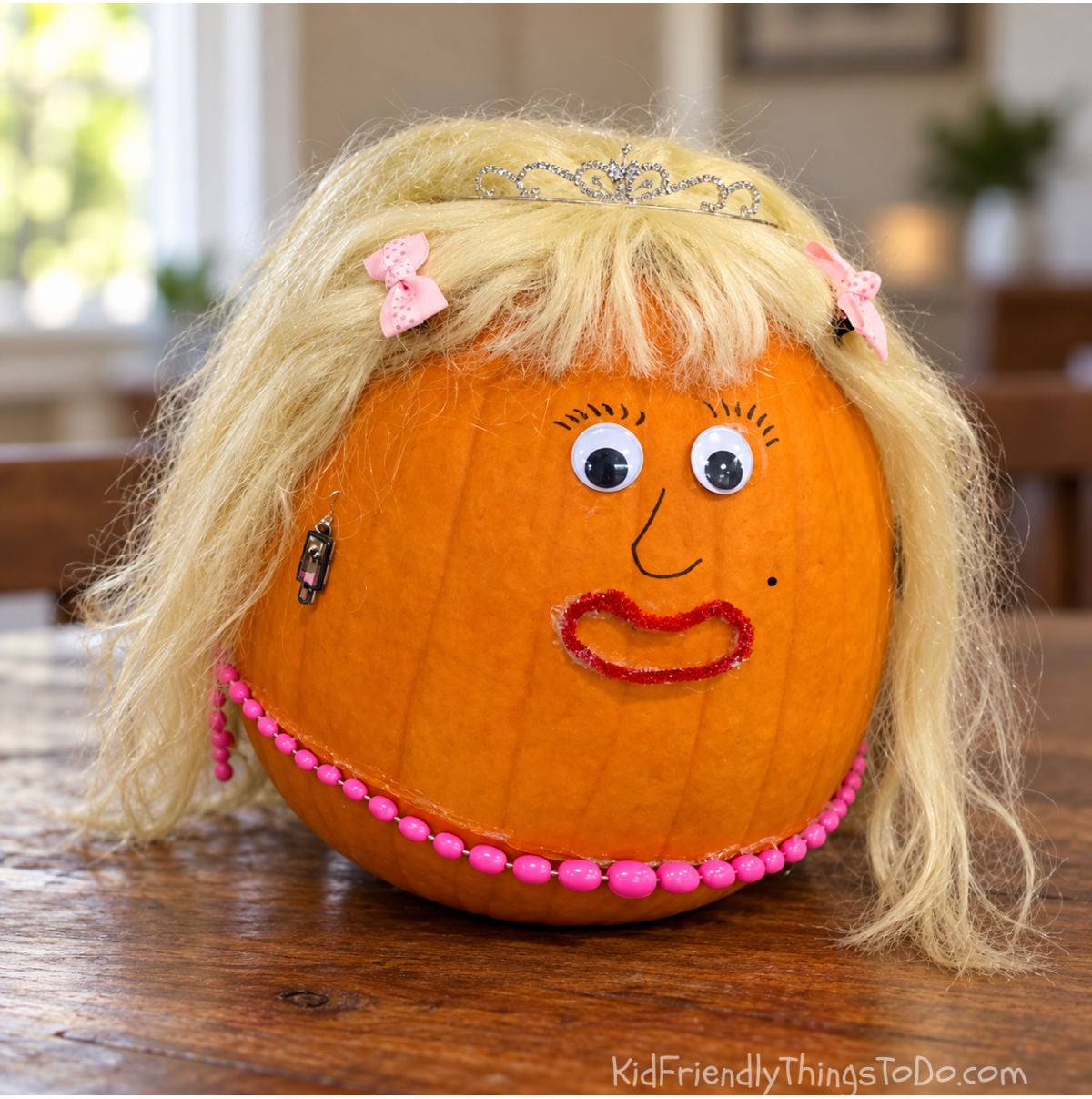Orange pumpkin decorated with a blonde wig, silver tiara, pink bows, googly eyes, red glitter lips, and a pink bead necklace.