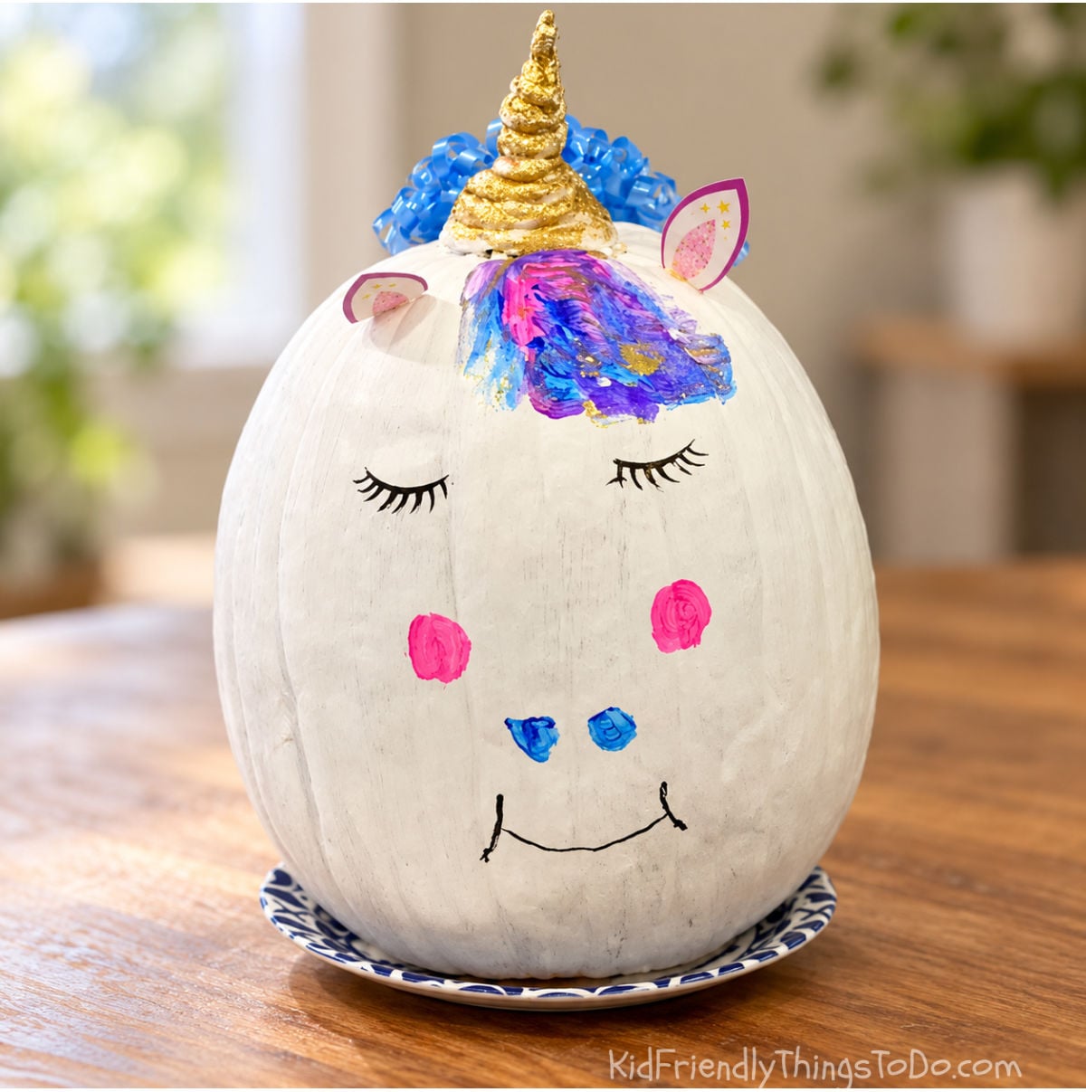 White unicorn pumpkin with a gold horn, blue ribbon curls, colorful painted forelock, closed eyelashes, and pink cheeks.