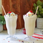 Milkshake recipe topped with whipped cream, rainbow sprinkles, and maraschino cherries in two old-fashioned soda glasses.