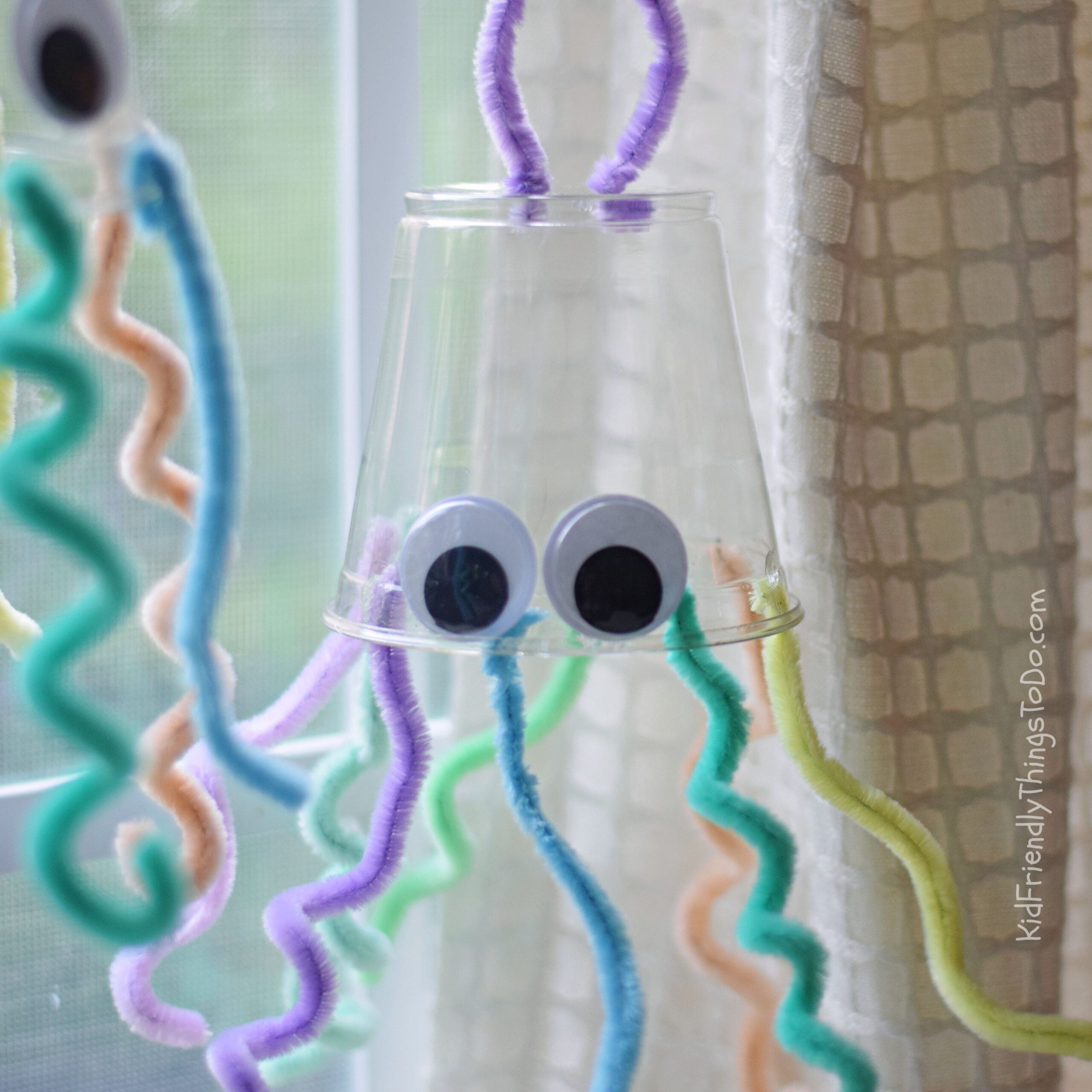 Plastic cup jellyfish craft hanging in a window with colorful pipe cleaner tentacles
