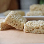Soft and gooey homemade Rice Krispie Treats stacked on a white plate in the background and on parchment paper in the front.