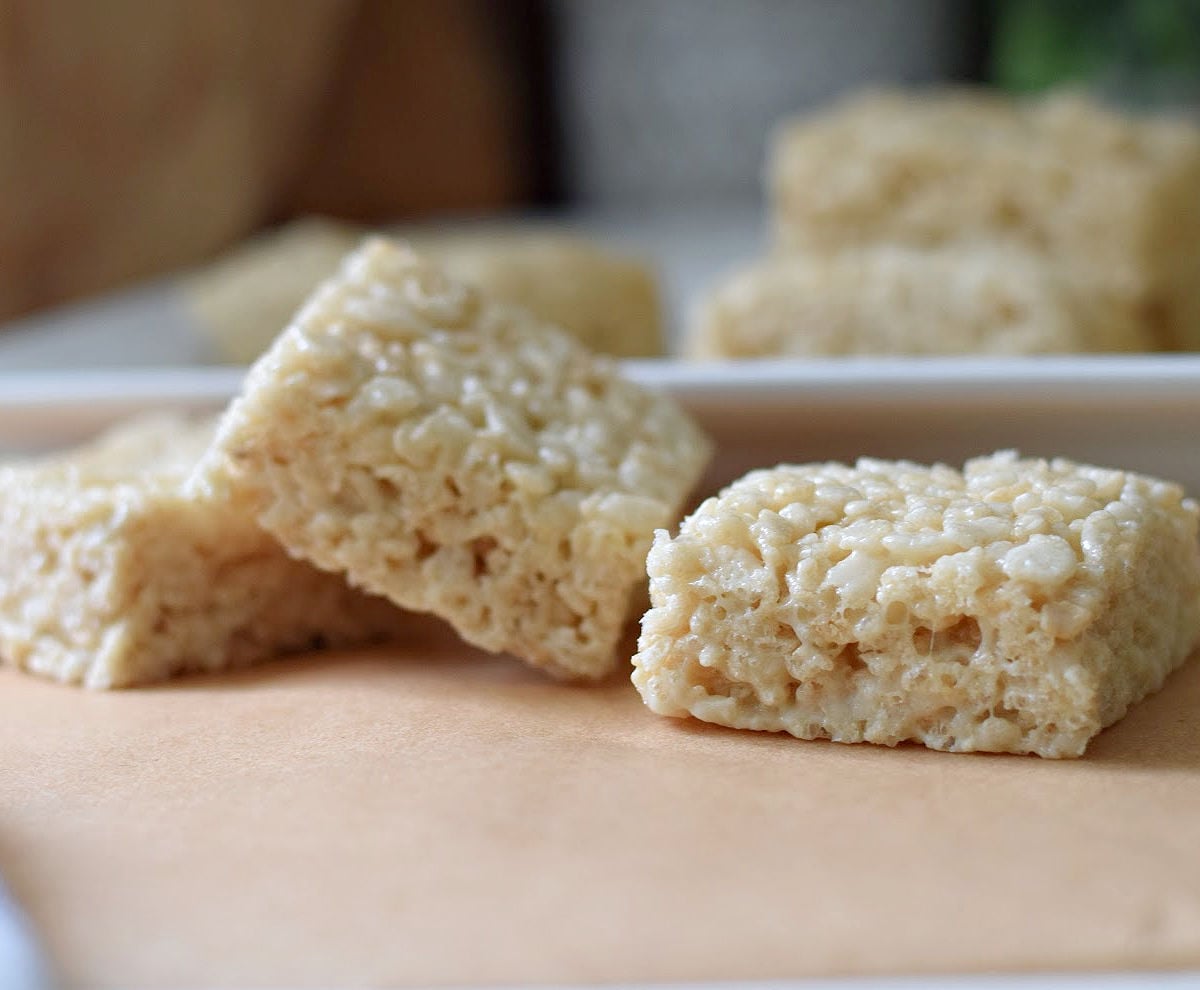 Soft and gooey homemade Rice Krispie Treats stacked on a white plate in the background and on parchment paper in the front.