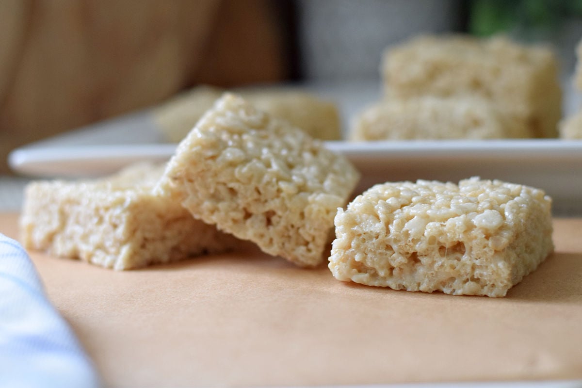 Soft and gooey homemade Rice Krispie Treats stacked on a white plate in the background and on parchment paper in the front.