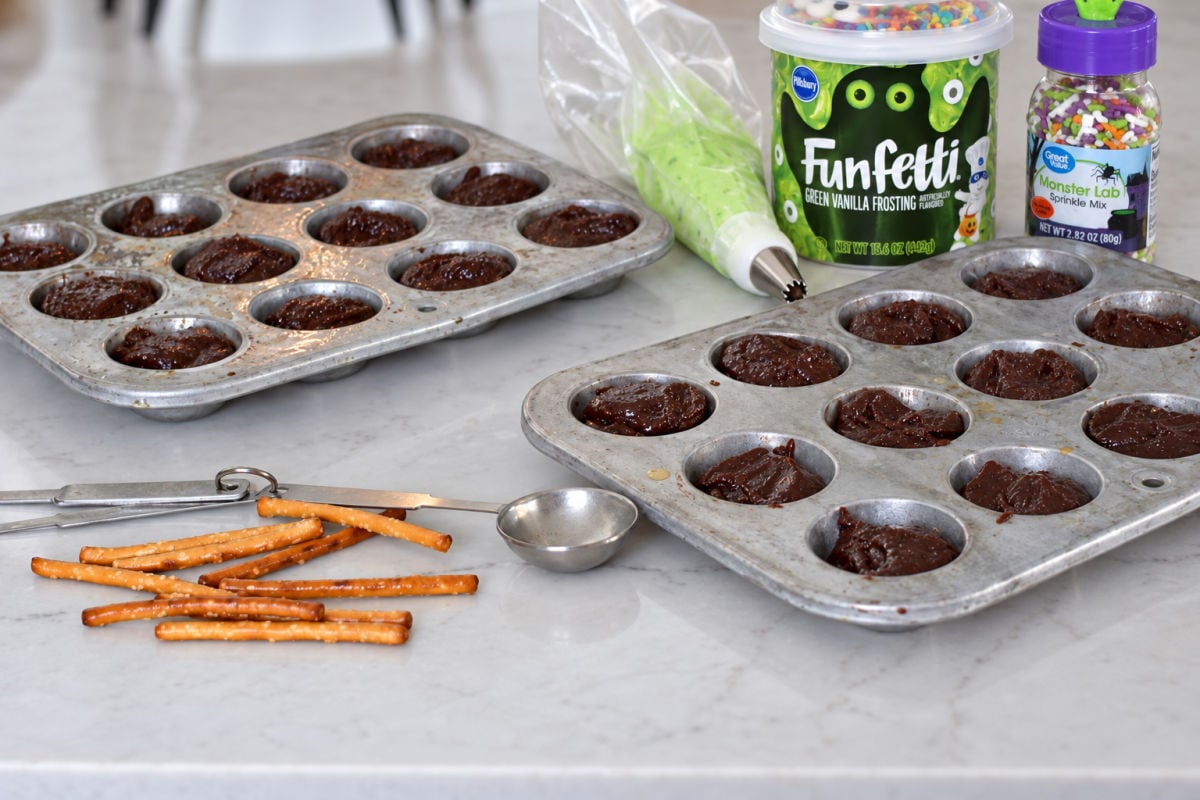 Ingredients for cauldron brownie bites, including brownie mix, green frosting, sprinkles, and pretzel sticks.