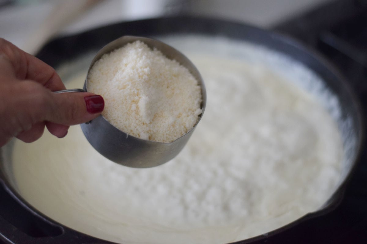 Hand holding a measuring cup of grated cheese above cream sauce in a skillet.