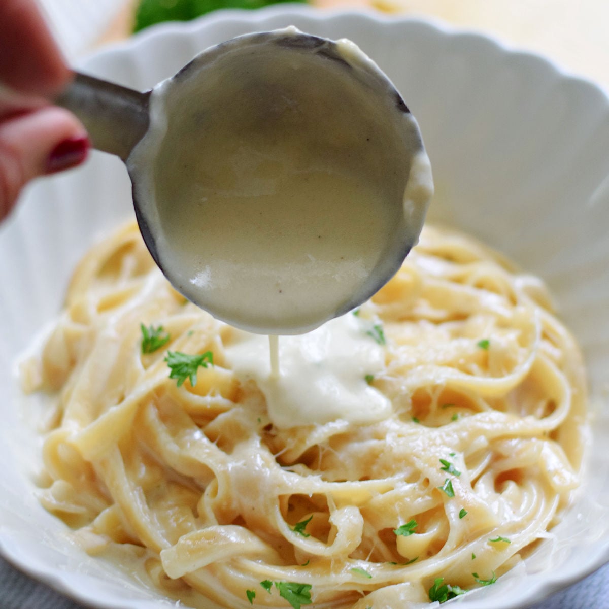 Alfredo sauce pouring from a metal ladle onto fettuccine topped with grated cheese and parsley in a white bowl.