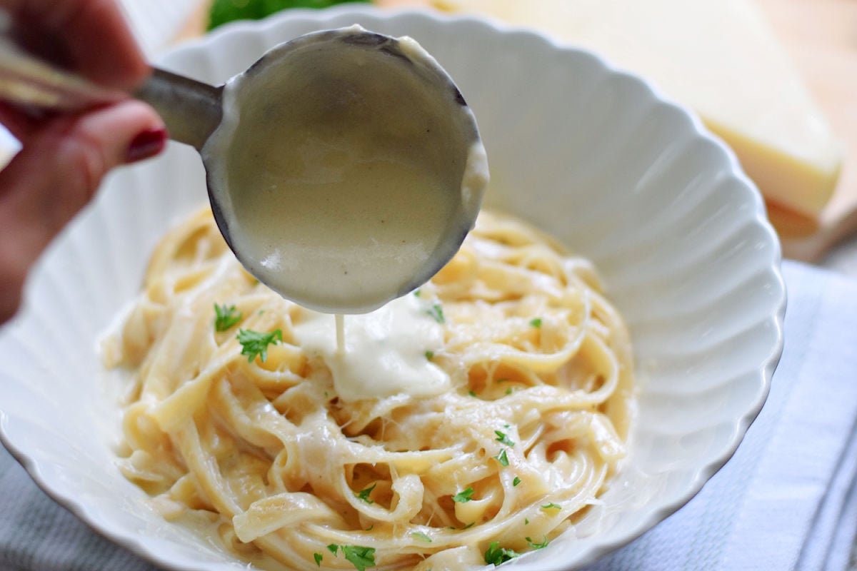 Alfredo sauce pouring from a metal ladle onto fettuccine topped with grated cheese and parsley in a white bowl.
