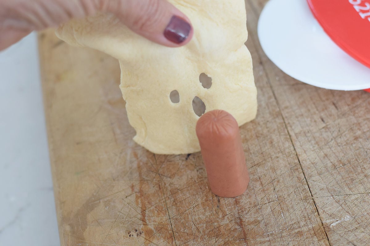 Lifting crescent dough with a cutout ghost face to drape over an upright hot dog half.