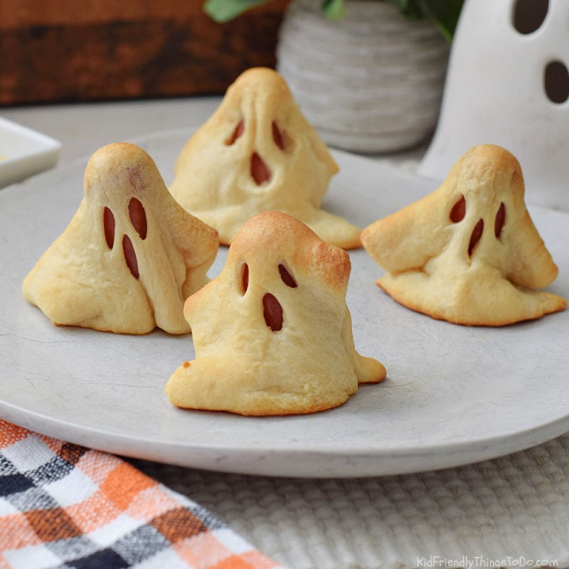 Four homemade Ghosts in a Blanket made with crescent dough and hot dogs, served on a white plate with mustard.