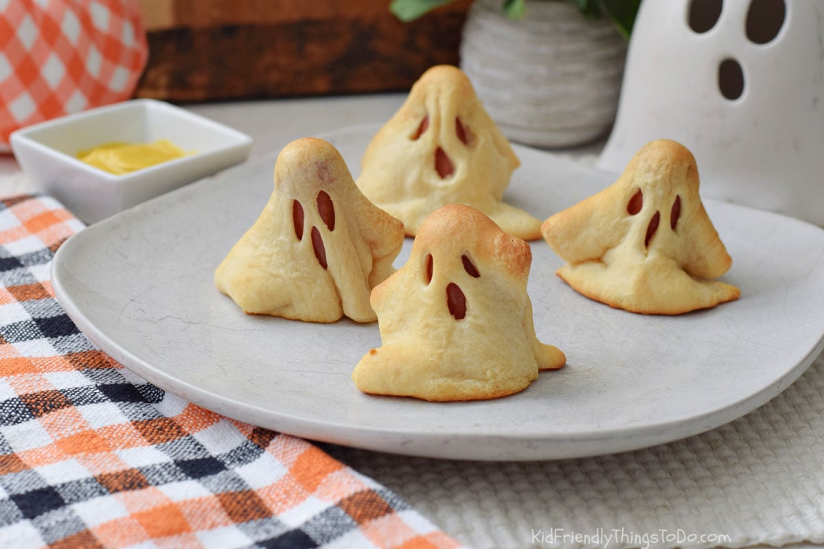 Four homemade Ghosts in a Blanket made with crescent dough and hot dogs, served on a white plate with mustard.