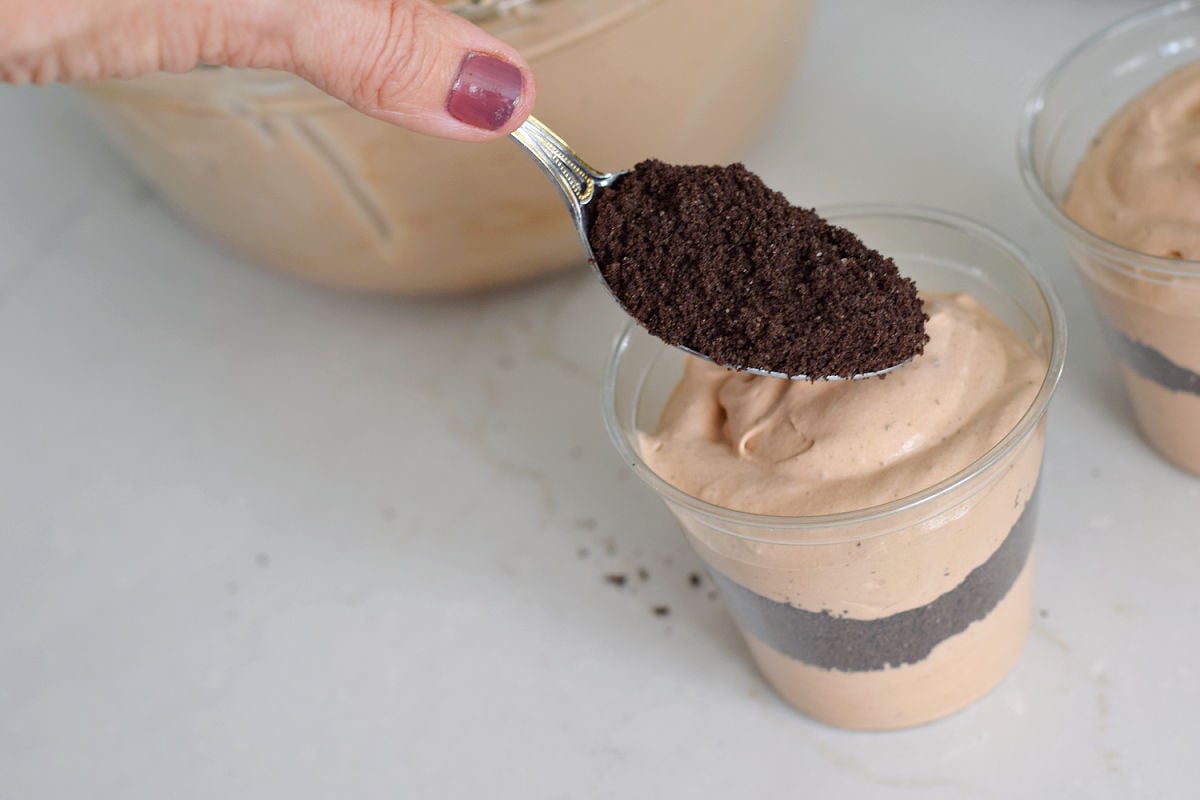 Spooning crushed Oreos over a dirt cup with alternating pudding and cookie layers.