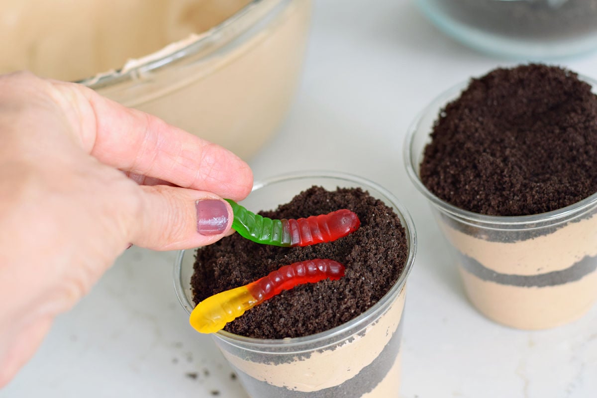 Adding gummy worms to the crushed Oreo topping on layered dirt pudding cups.