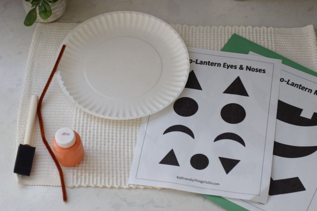 Supplies for a paper plate pumpkin craft, including printable jack o lantern face sheets, paint, a foam brush, paper, and a pipe cleaner.
