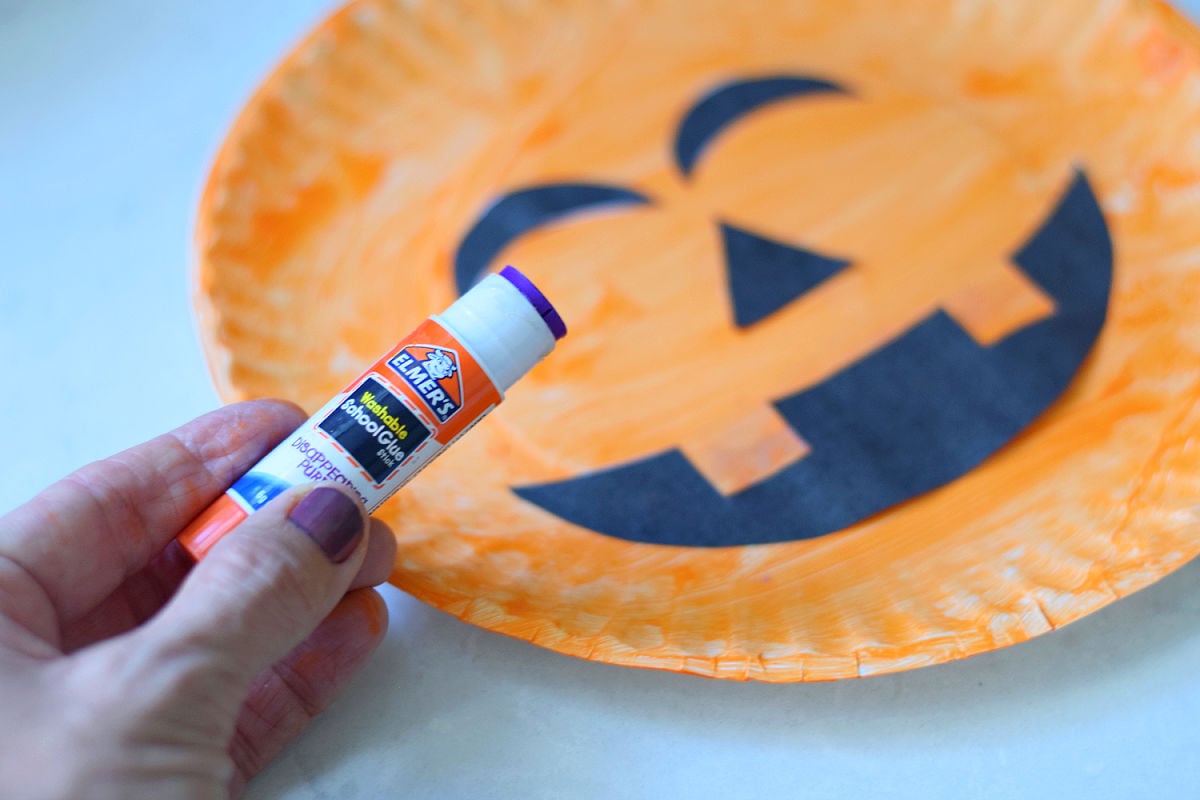 Black jack o lantern face cutouts arranged on an orange paper plate beside a glue stick.