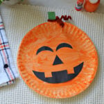 Orange paper plate jack o lantern with black printable face cutouts, a green paper stem, and a brown pipe cleaner curl.