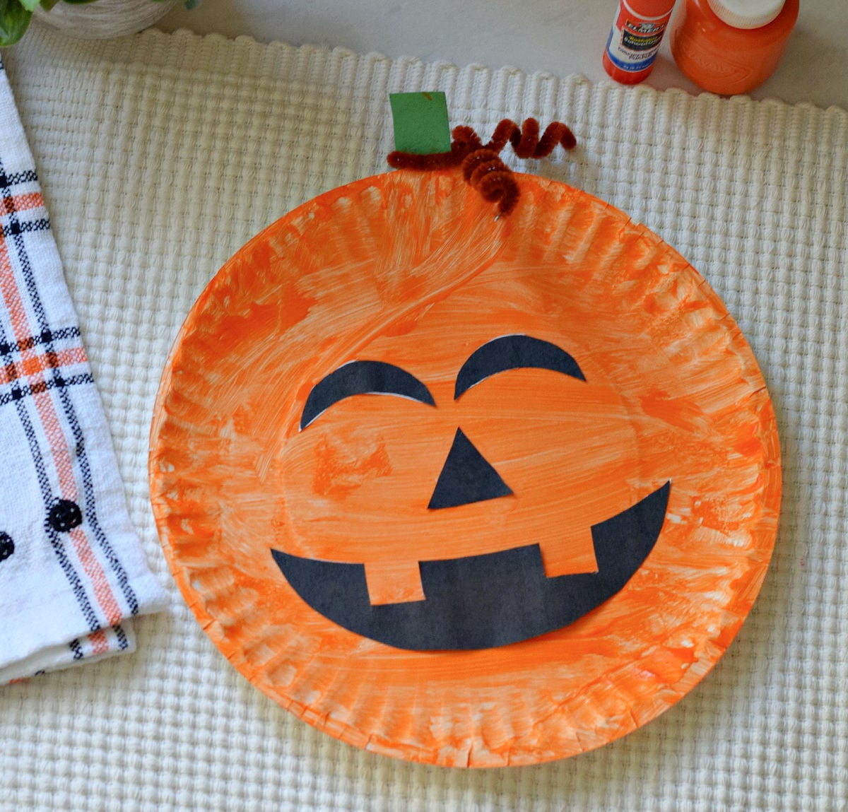 Orange paper plate jack o lantern with black printable face cutouts, a green paper stem, and a brown pipe cleaner curl.