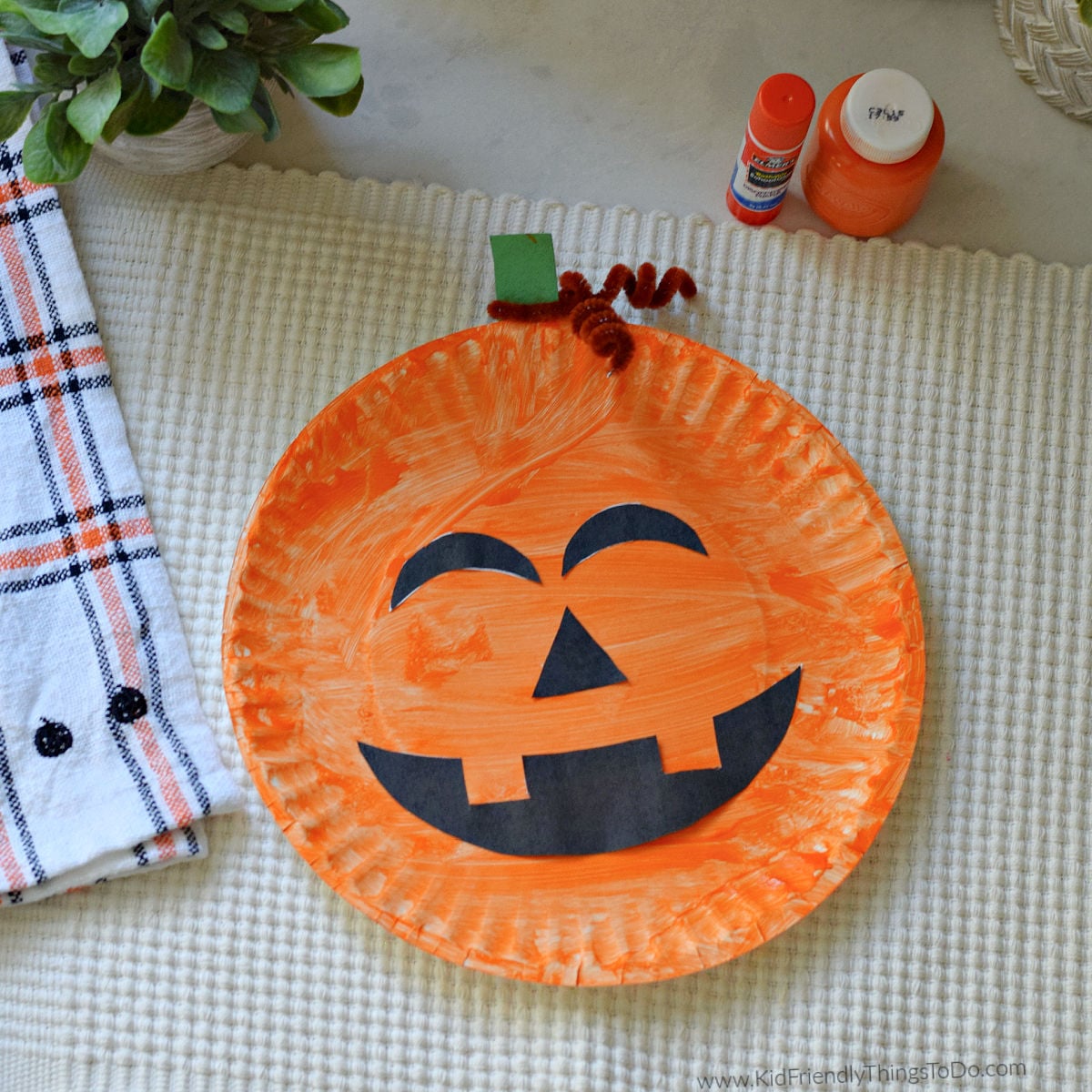 Orange paper plate jack o lantern with black printable face cutouts, a green paper stem, and a brown pipe cleaner curl.