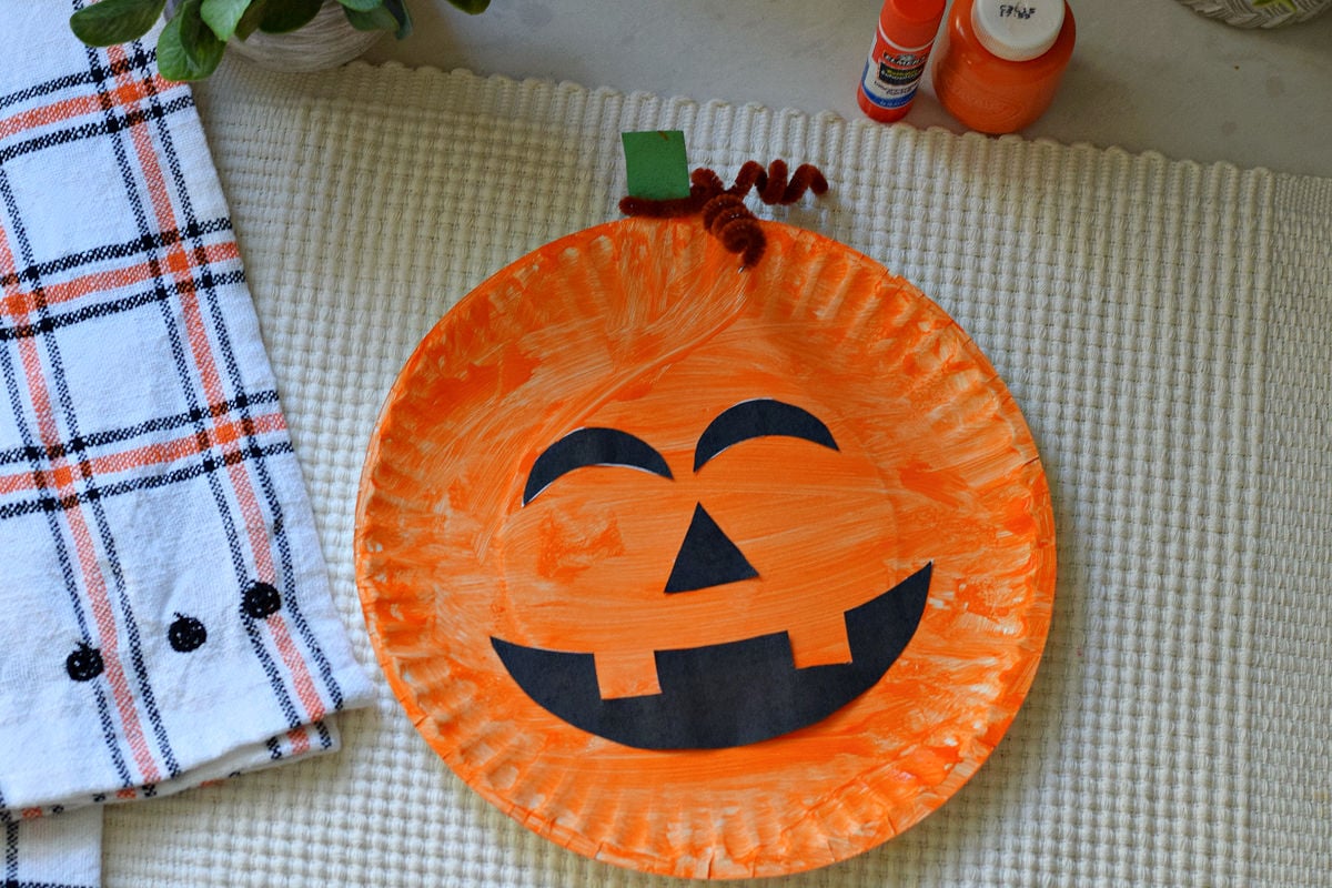 Orange paper plate jack o lantern with black printable face cutouts, a green paper stem, and a brown pipe cleaner curl.