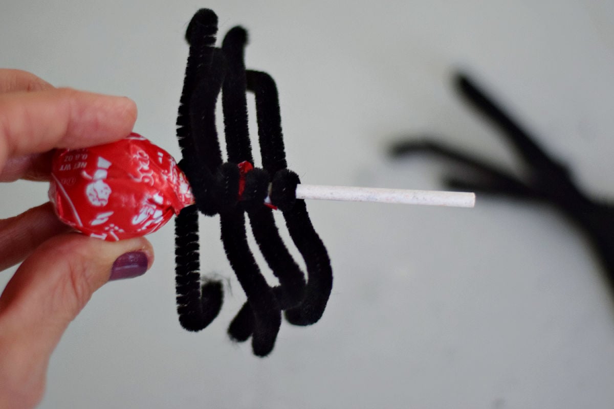 Hand holding a wrapped Tootsie Pop with black pipe cleaners wrapped around the stick and bent into spider legs.