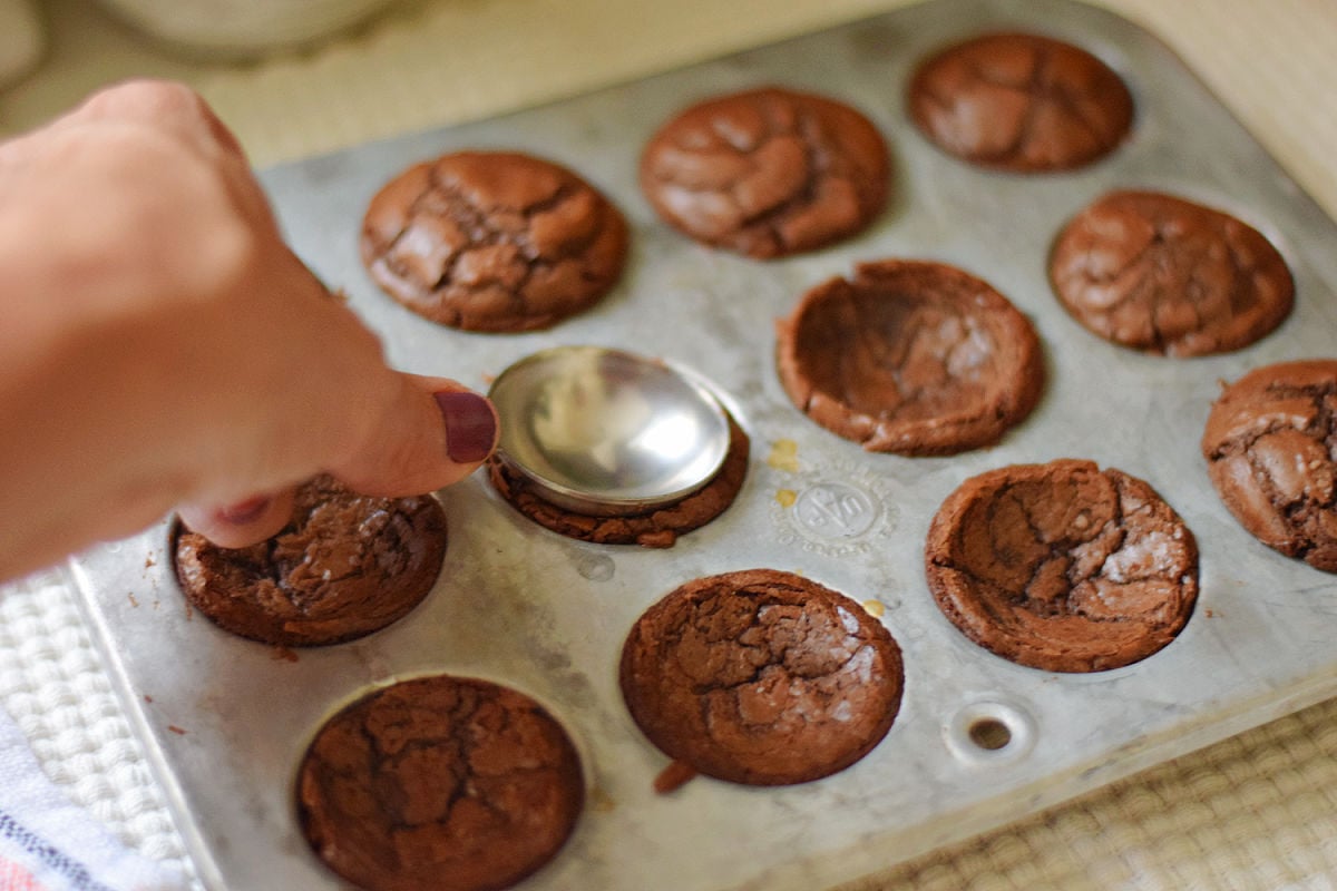 Rounded back of a measuring spoon pressing a shallow bowl into a warm mini brownie.