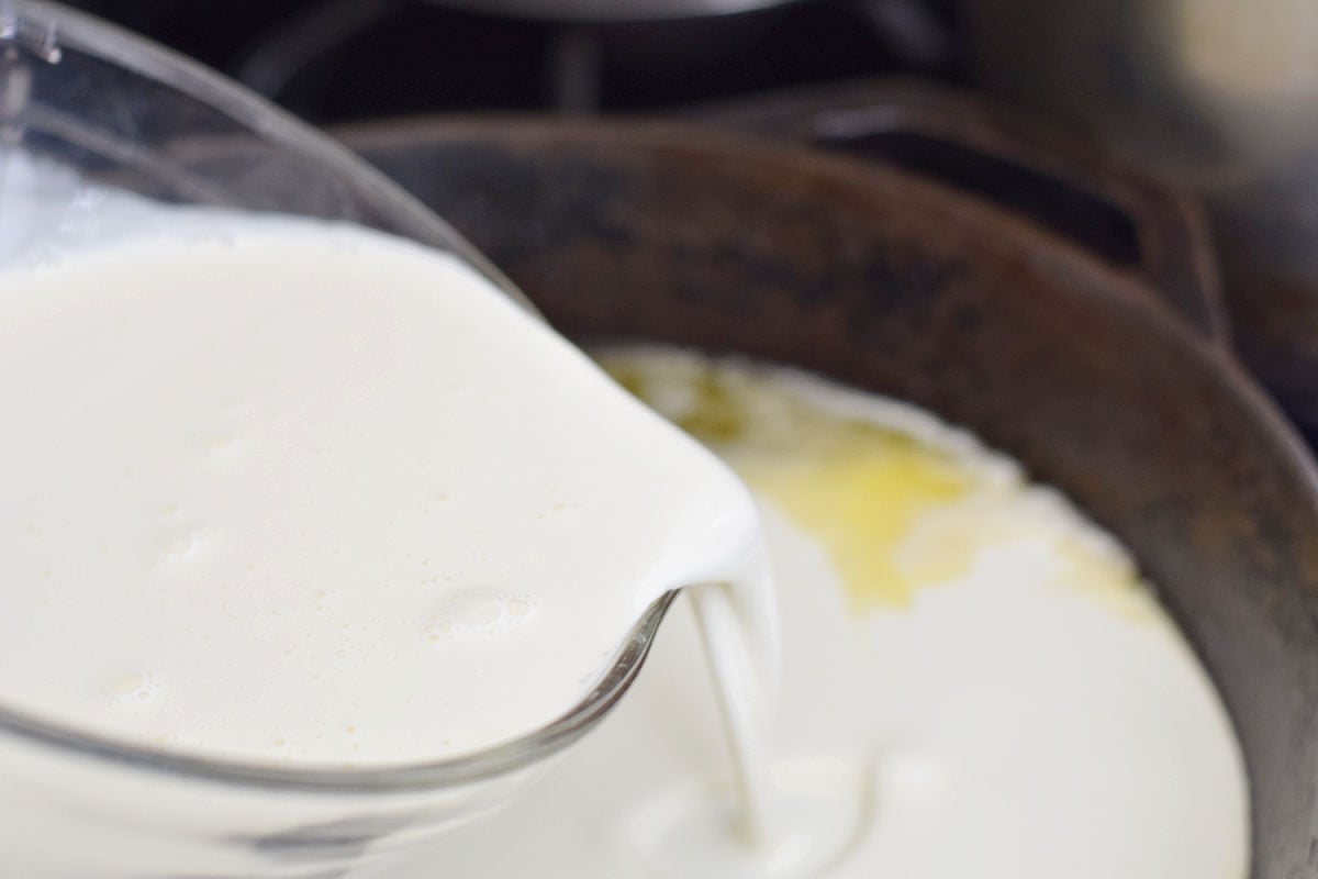 Heavy cream pouring from a glass measuring cup into a skillet with melted butter.